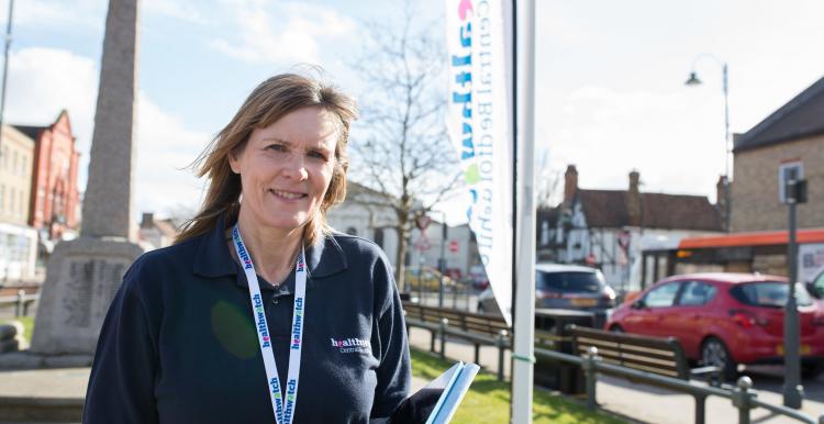 Female volunteer wearing a Healthwatch t shirt and lanyard standing in front of a flag